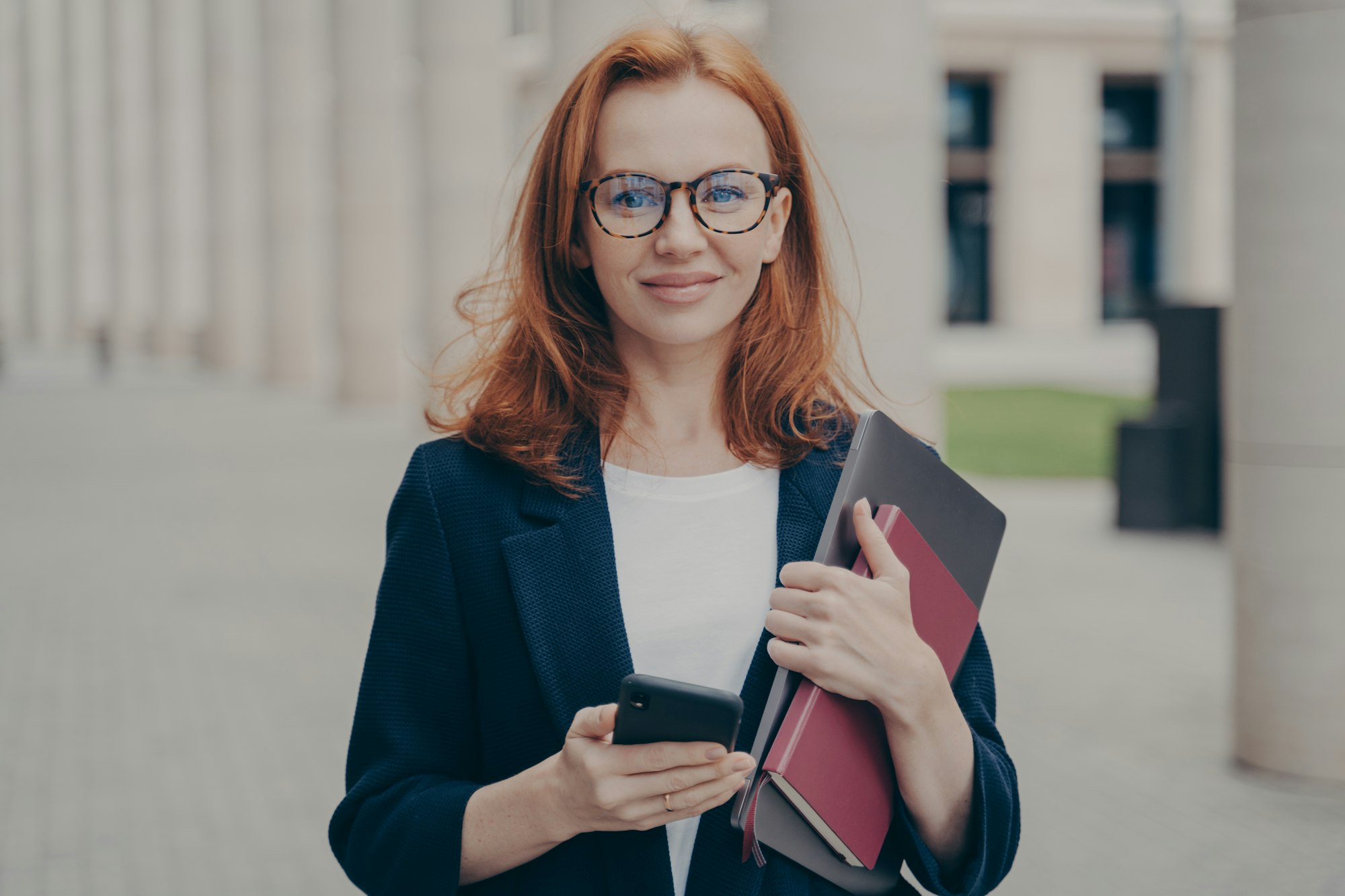 Confident beautiful red-haired female business consultant holding modern smartphone and laptop